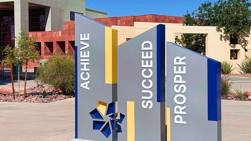 The Achieve, Succeed, Prosper sculpture stands outside a CSN campus building, featuring three vertical panels with the words “Achieve,” “Succeed,” and “Prosper” alongside the college logo.