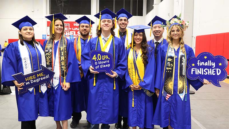Group of graduating students in blue caps and gowns smiling and holding celebratory signs at an indoor commencement event.
