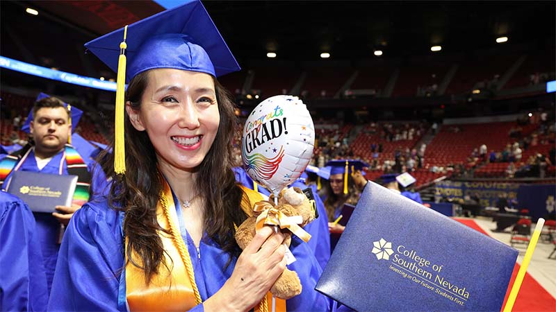 Smiling graduate in a blue cap and gown holds a diploma folder and a small teddy bear with a 'Congrats Grad' balloon while standing inside a commencement arena.