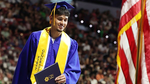 Graduate in a blue cap and gown wearing a gold honor stole, holding a diploma folder, standing near an American flag at a commencement ceremony.