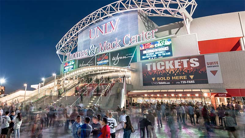 Exterior view of the Thomas & Mack Center at UNLV at night, with illuminated signage above the main entrance and crowds of people gathered on the steps and plaza outside.