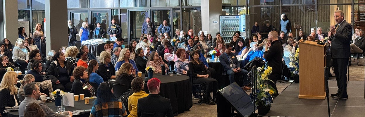 College of Southern Nevada President Dr. Klippenstein speaks at a podium during Convocation as faculty and staff gather around tables in a large atrium, listening to the presentation. An American Sign Language interpreter stands beside the stage, and floral arrangements decorate the podium.