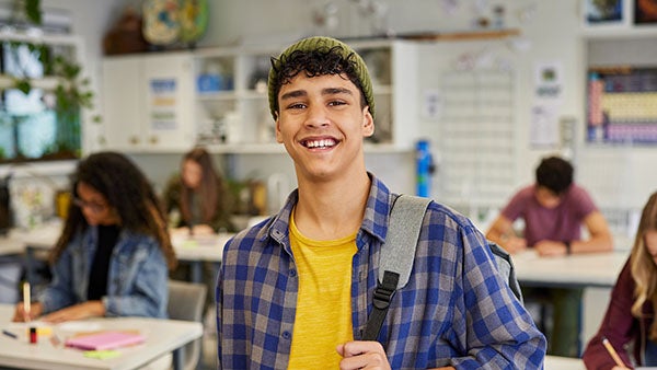 Smiling student wearing a backpack standing in a classroom while other students work at desks in the background.
