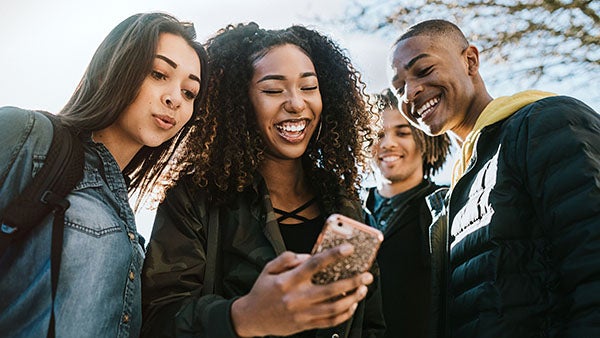 Group of college-age students outdoors smiling and looking at a smartphone together.