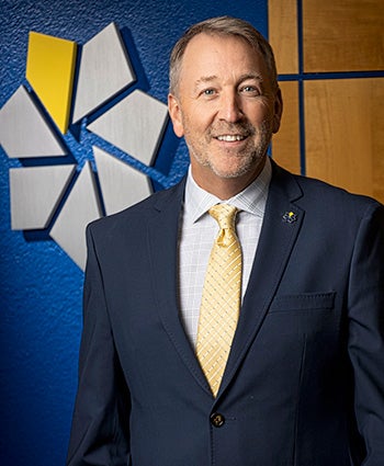 Official portrait of College of Southern Nevada President Dr. Stacy Klippenstein wearing a navy suit, white shirt, and yellow tie against a gray background.