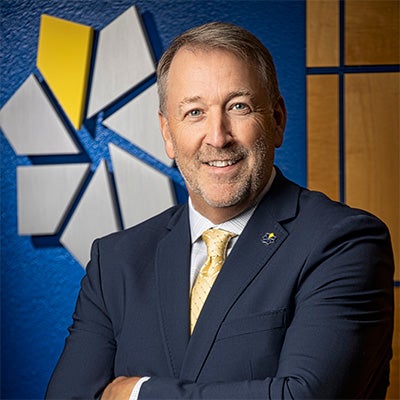 Portrait of College of Southern Nevada President Dr. Stacy Klippenstein smiling with arms crossed in front of the CSN logo.