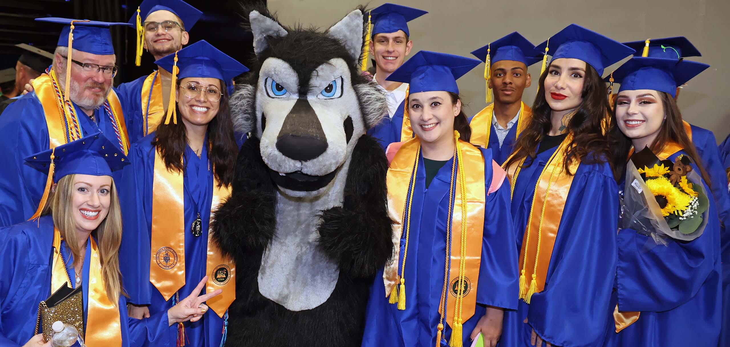 A group of College of Southern Nevada graduates in blue caps and gowns pose together with the CSN mascot during commencement.