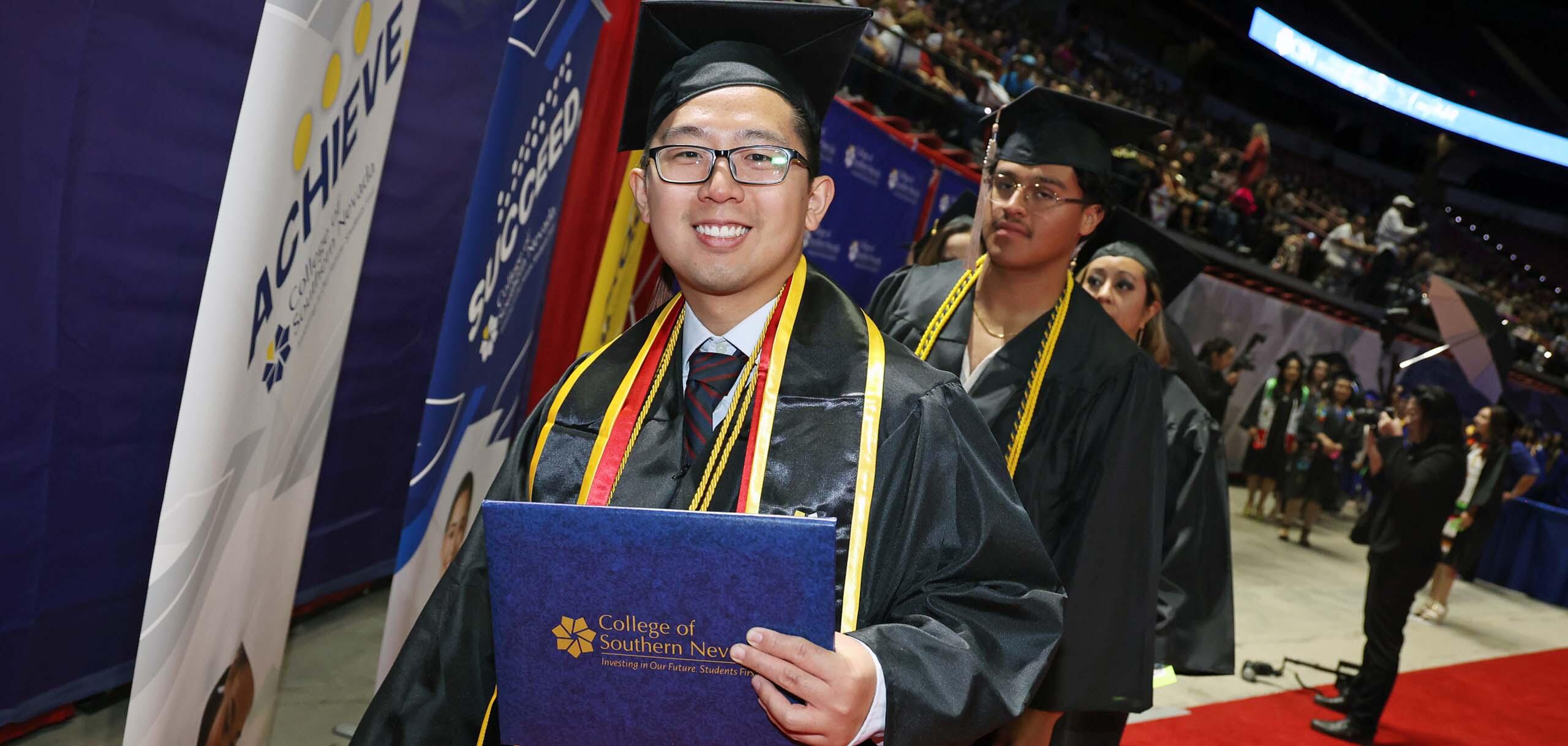 A CSN graduate in cap and gown holds a diploma while processing during a commencement ceremony.
