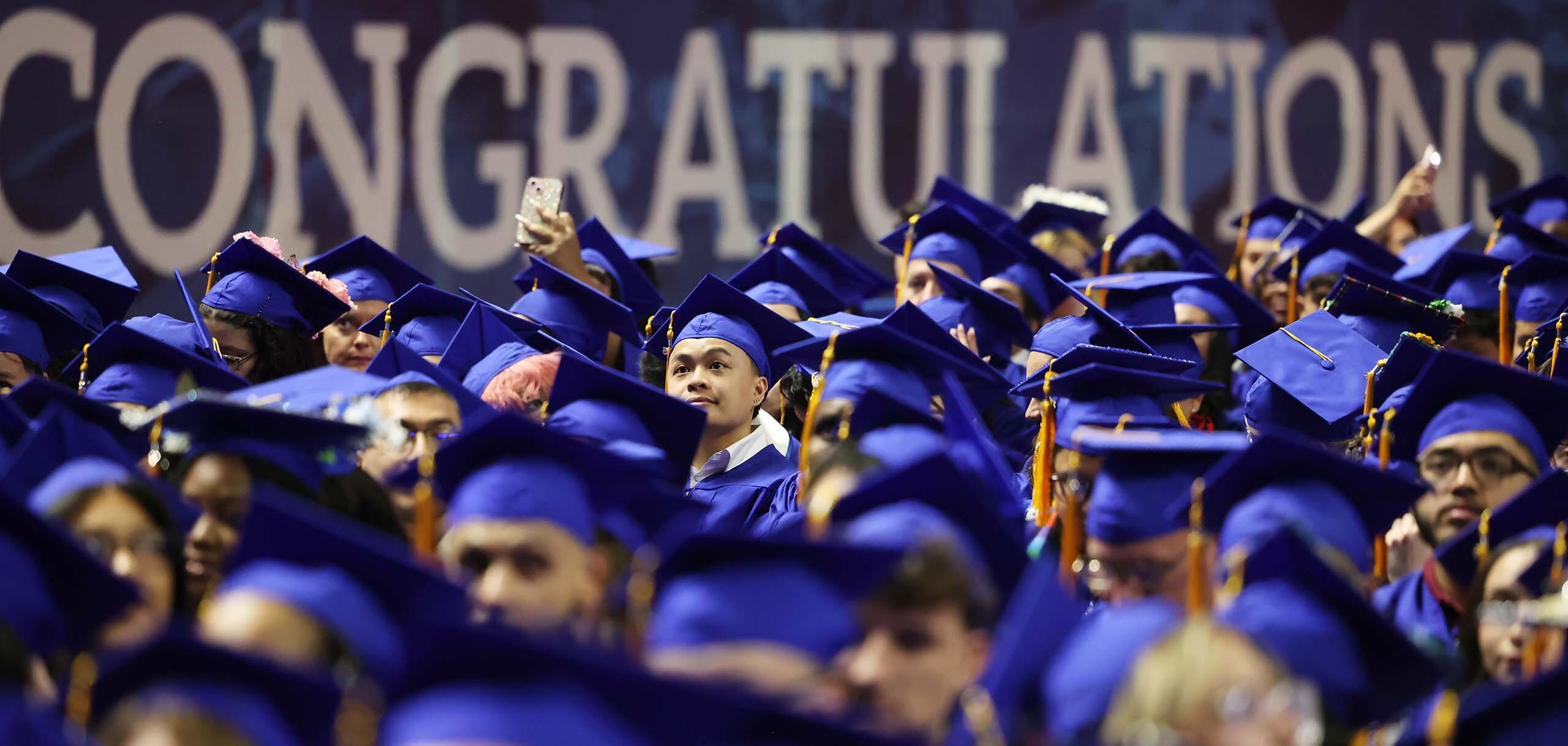 College of Southern Nevada graduates in blue caps and gowns gathered at a commencement ceremony with a large ‘Congratulations’ sign in the background.
