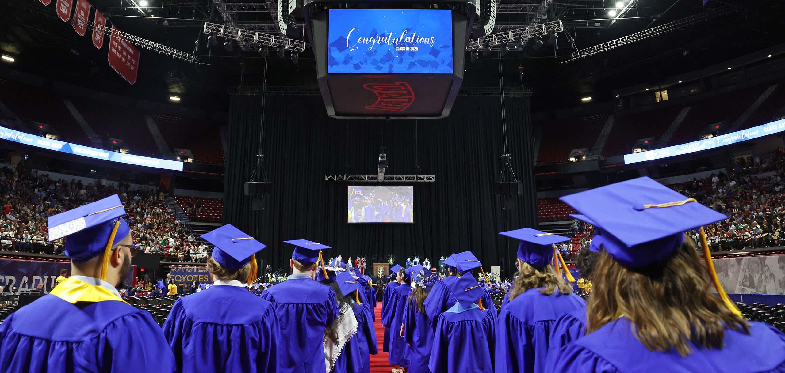 CSN graduates in caps and gowns process toward the stage during a commencement ceremony in a crowded arena.