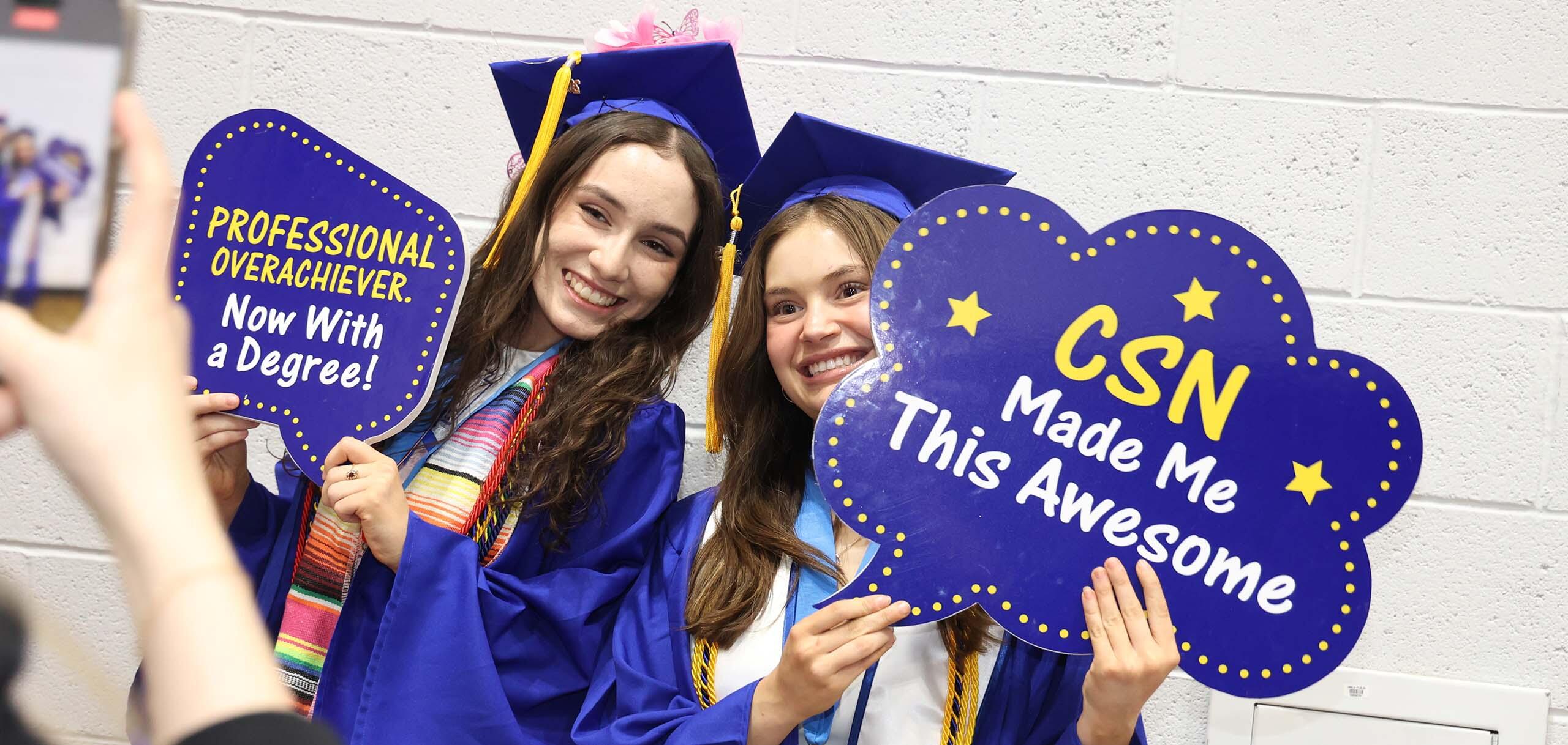 Two graduates in caps and gowns smile while holding signs celebrating their achievements at CSN.”