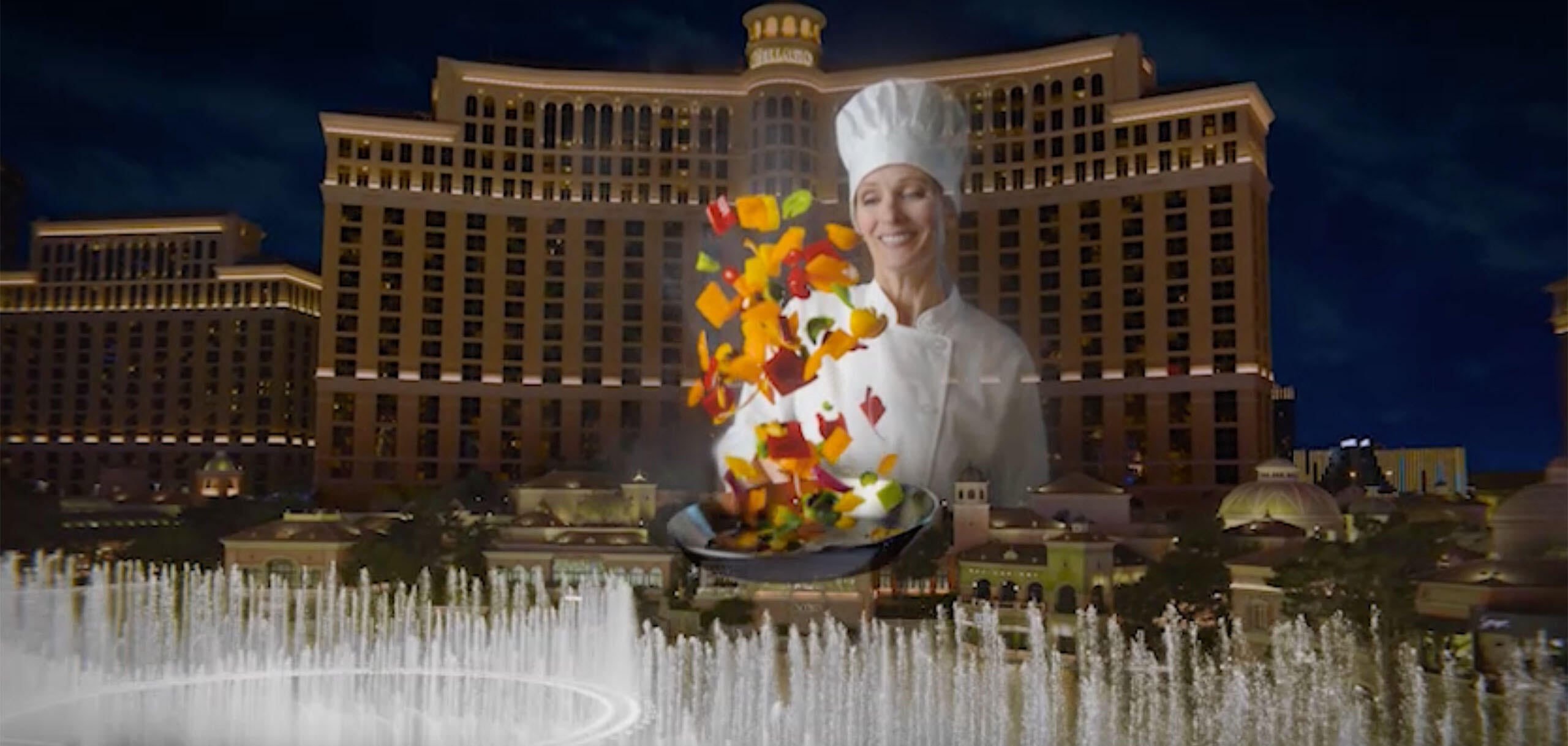 A chef tosses colorful vegetables in a pan in front of the Bellagio resort and fountains on the Las Vegas Strip at night.