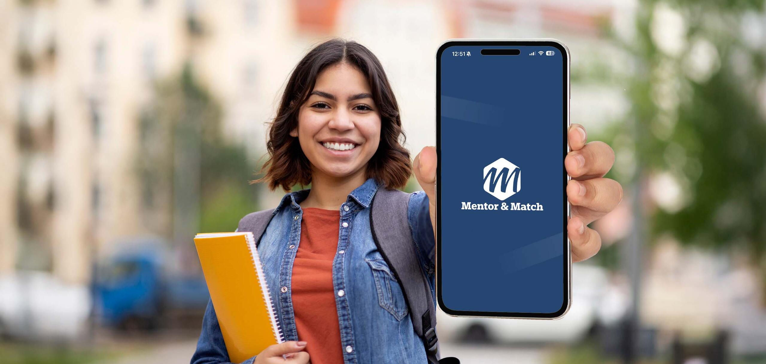 Smiling student outdoors holding a smartphone displaying the Mentor & Match app, while carrying a backpack and a yellow notebook.